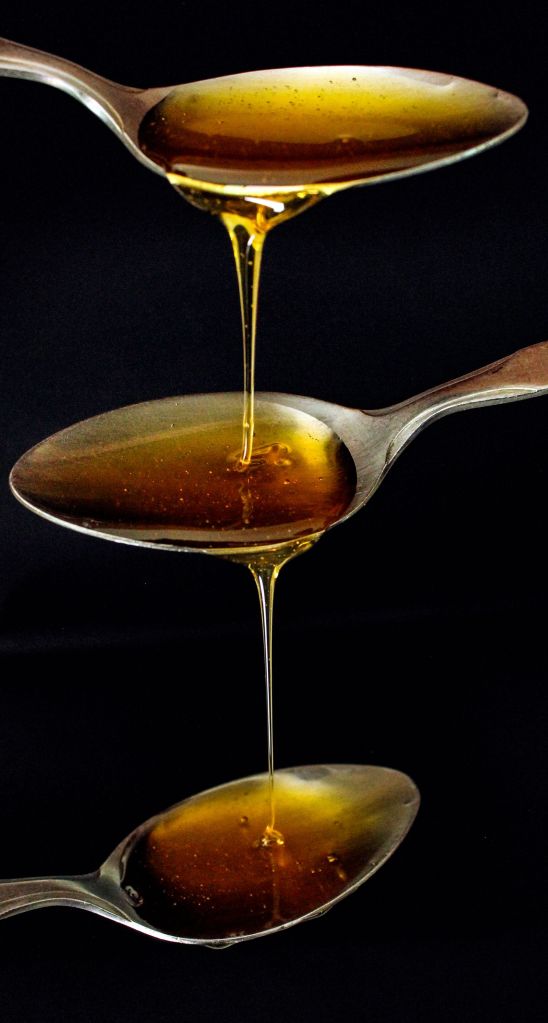 Thick golden syrup flowing from three spoons on a black background