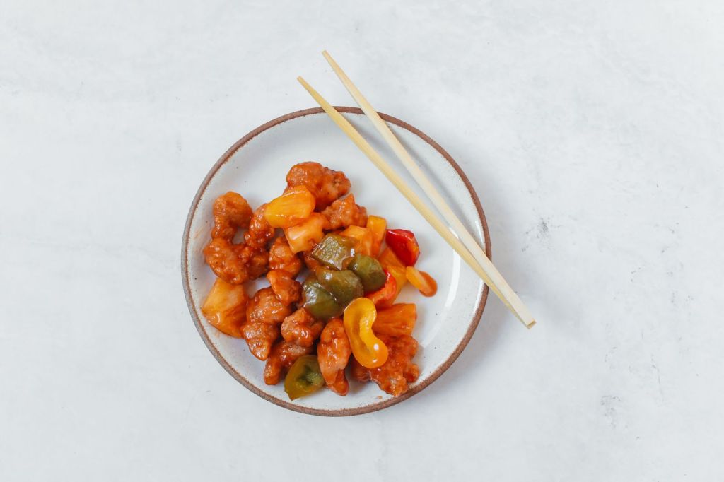 Crispy sweet and sour pork with vegetables and glossy sauce in a white bowl with wooden chopsticks